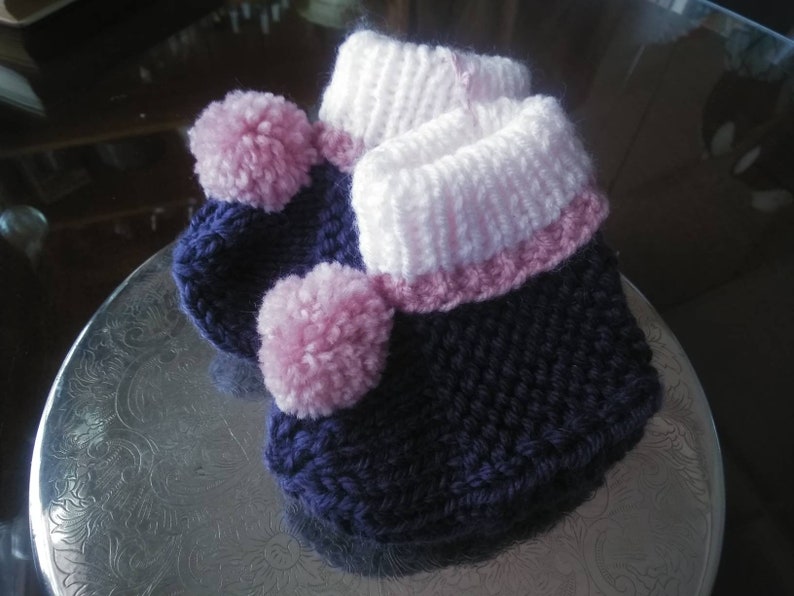 May include: A pair of knitted baby booties with pink and purple yarn. The booties have a pink pom-pom on the top.