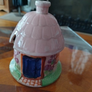 May include: A pink ceramic cottage-shaped sugar bowl with a blue door and floral details. The bowl has a thatched roof and a chimney.