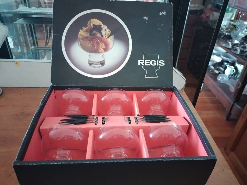 Puede incluir: Una caja negra con interior rojo contiene seis vasos de vidrio transparente. La caja tiene la palabra "REGIS" impresa en la parte delantera. Los vasos est&aacute;n dispuestos en dos filas de tres. La caja est&aacute; abierta y los vasos son visibles.