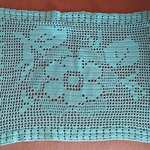 May include: A rectangular, light turquoise crocheted doily with a floral design. The openwork pattern features a large central flower and surrounding floral elements. The doily is made of a soft, textured yarn.