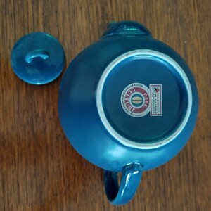 Puede incluir: Una tetera de cer&aacute;mica azul con tapa y asa. La tetera tiene un c&iacute;rculo blanco en la parte inferior con el texto "Cafe Culture" y "Microwave and Dishwasher Safe Porcelain".