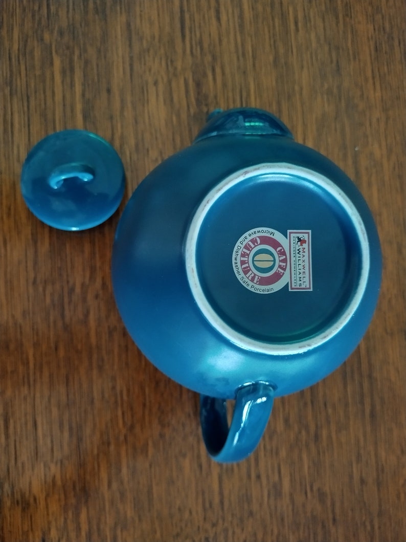 Puede incluir: Una tetera de cer&aacute;mica azul con tapa y asa. La tetera tiene un c&iacute;rculo blanco en la parte inferior con el texto "Cafe Culture" y "Microwave and Dishwasher Safe Porcelain".