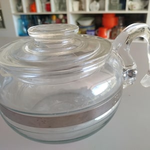 May include: A clear glass teapot with a lid and a curved handle. The teapot has a silver band around the middle. The teapot is designed for serving tea and other hot beverages. The teapot is made of glass.