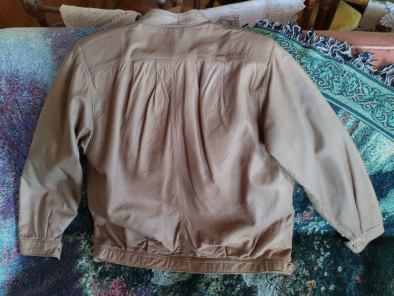 Puede incluir: Una chaqueta de cuero marr&oacute;n claro con cierre de cremallera y cuello abotonado. La chaqueta tiene una textura lisa y desgastada y una apariencia ligeramente arrugada.