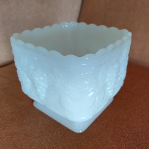 May include: A square, white milk glass bowl with a scalloped rim. The sides of the bowl are decorated with a raised floral pattern. The bowl sits on a square base. This vintage piece is suitable for home decor.