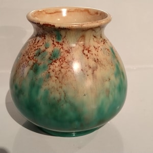 May include: A small, bulbous ceramic vase featuring a smooth, glossy finish. The glaze displays a blended gradient of deep teal and turquoise at the base, transitioning into a creamy off-white and speckled brown pattern toward the flared rim.
