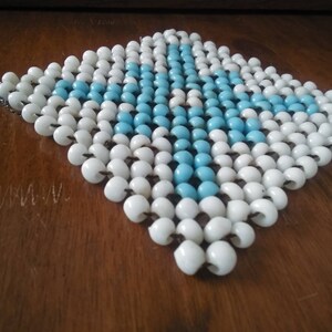 May include: A square-shaped beaded coaster with a blue and white pattern. The coaster is made of small, round beads that are strung together.