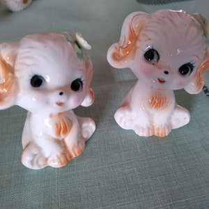 May include: Two white ceramic figurines of dogs with large eyes, brown ears, and green flower accents. The dogs are sitting with their paws tucked in.