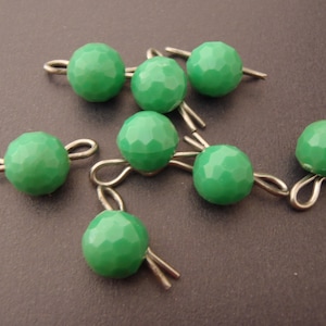 May include: A group of eight green faceted glass beads with silver metal loops. The beads are arranged in a cluster on a grey surface.