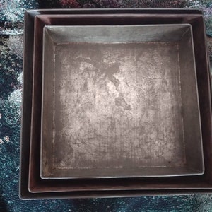 May include: Three nested metal baking pans, all square shaped, with a silver finish. The pans are slightly worn and have a rustic look.