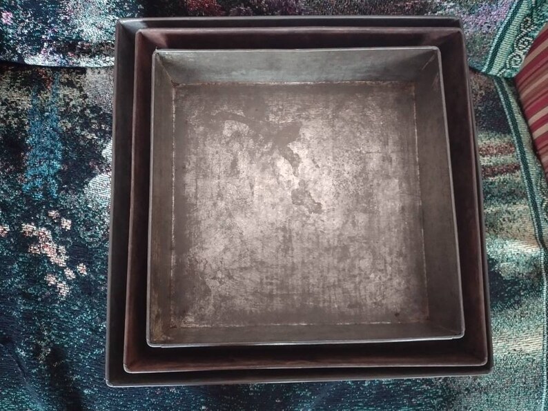 May include: Three nested metal baking pans, all square shaped, with a silver finish. The pans are slightly worn and have a rustic look.