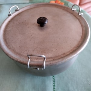 May include: A round, aluminum cooking pot with a lid. The pot has two metal handles and a black knob on the lid. The pot is sitting on a light green tablecloth.