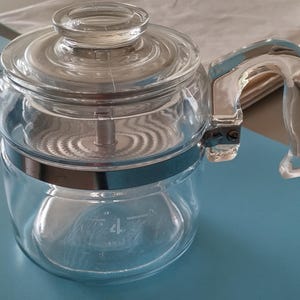 May include: A clear glass coffee percolator with a clear lid and handle. The percolator has a silver band around the middle and a measurement indicator for 4 cups. The handle is clear and attached to the side.