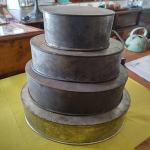 K&ouml;nnte beinhalten: Ein Stapel von vier runden, vintage Kuchenformen aus Metall in verschiedenen Gr&ouml;&szlig;en. Die Formen haben eine silber-graue Oberfl&auml;che mit Gebrauchsspuren. Die gr&ouml;&szlig;te Form hat einen goldfarbenen Boden. Die Formen sind in einer gestaffelten Anordnung angeordnet.
