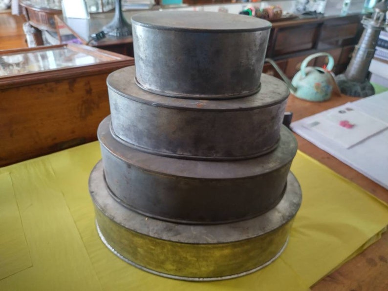 K&ouml;nnte beinhalten: Ein Stapel von vier runden, vintage Kuchenformen aus Metall in verschiedenen Gr&ouml;&szlig;en. Die Formen haben eine silber-graue Oberfl&auml;che mit Gebrauchsspuren. Die gr&ouml;&szlig;te Form hat einen goldfarbenen Boden. Die Formen sind in einer gestaffelten Anordnung angeordnet.