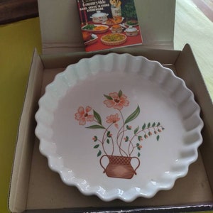 以下が含まれることがあります： 白いセラミック製のパイ皿で、縁が波打っていて、ピンクの花と緑の葉が茶色の壺に入っている花柄のデザインが施されています。皿は、箱に入っていて、箱には「Countryside Bake, Serve 'n Store Stoneware」と書いてあります。