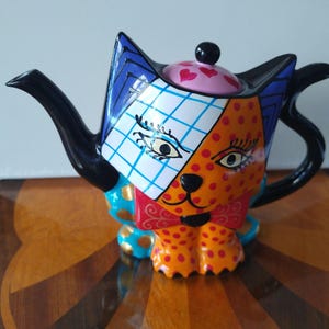 May include: A ceramic teapot shaped like a cat with a vibrant, multi-colored design. The cat features a white grid pattern on one side of its face, orange with red polka dots on the other, a red bow tie, and a pink lid decorated with red hearts.