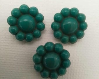 Può includere: Tre bottoni a forma di fiore color teal. Ogni bottone ha un cerchio centrale in rilievo circondato da piccoli petali arrotondati. I bottoni sono realizzati in materiale lucido e sono disposti su una superficie bianca.