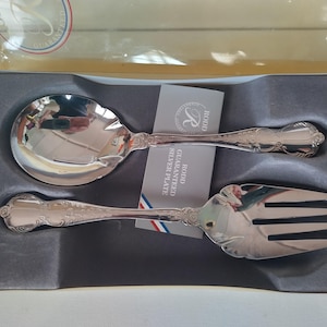 May include: A silver-plated serving spoon and fork set with ornate embossed handles, presented in a gray molded tray inside a box. A small card between the utensils reads Rodd Guaranteed Silver Plate with a circular logo and a small flag graphic.