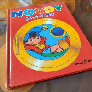 以下が含まれることがあります： 黄色い文字で「Noddy goes to sea」というタイトルが書かれた赤い本。表紙には、赤いシャツと青い帽子をかぶったノッディーが丸窓の中にいるという漫画のイラストが描かれており、背景は黄色です。