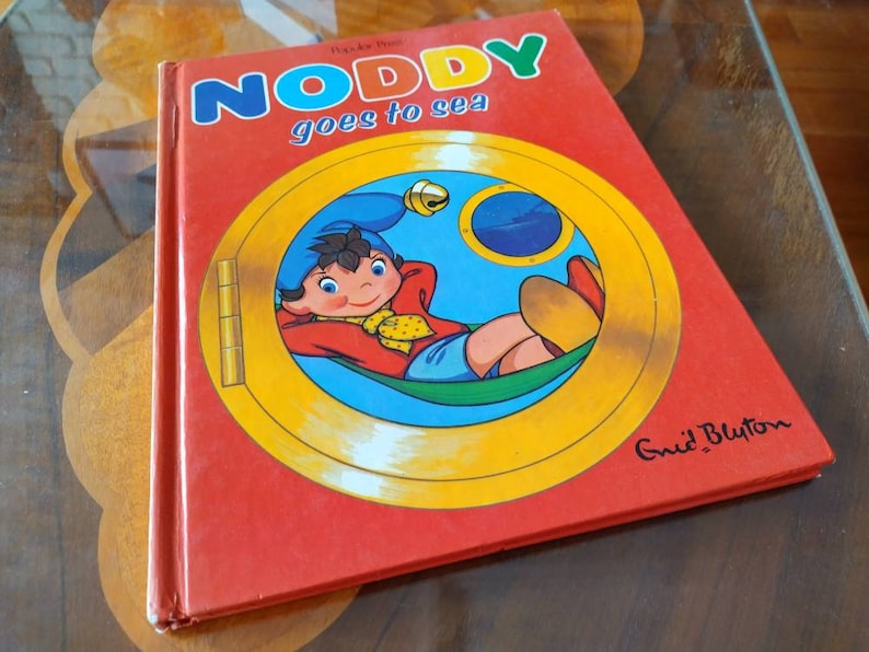 以下が含まれることがあります： 黄色い文字で「Noddy goes to sea」というタイトルが書かれた赤い本。表紙には、赤いシャツと青い帽子をかぶったノッディーが丸窓の中にいるという漫画のイラストが描かれており、背景は黄色です。