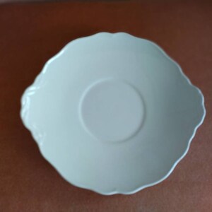 May include: A white ceramic saucer with a scalloped edge and a slightly raised center.