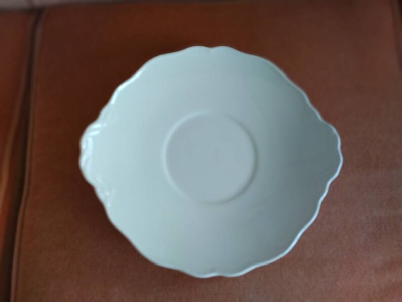 May include: A white ceramic saucer with a scalloped edge and a slightly raised center.