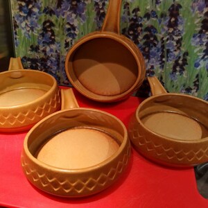May include: Four brown ceramic ramekins with handles. The ramekins have a diamond pattern around the rim. The ramekins are on a red surface.