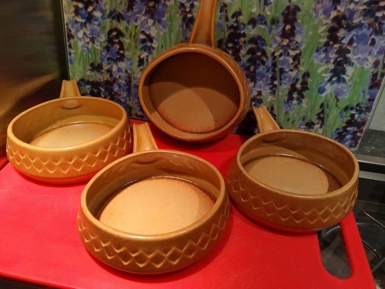 May include: Four brown ceramic ramekins with handles. The ramekins have a diamond pattern around the rim. The ramekins are on a red surface.