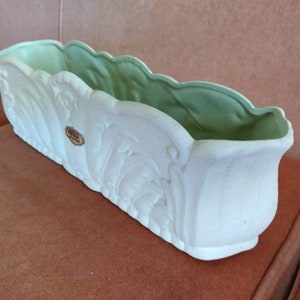 May include: A rectangular, white ceramic planter with a scalloped rim and embossed floral designs. The interior is a light green colour. A small, round sticker is affixed to the front. This decorative planter is suitable for indoor or outdoor use.