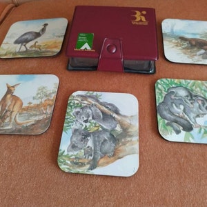 May include: Set of six square coasters featuring Australian animals. The coasters are made of wood and have a glossy finish. Each coaster features a different animal, including a kangaroo, a koala, a possum, an echidna, a platypus, and an emu. The coasters are packaged in a red box with the words "Royal Australian Mint" printed on the front.