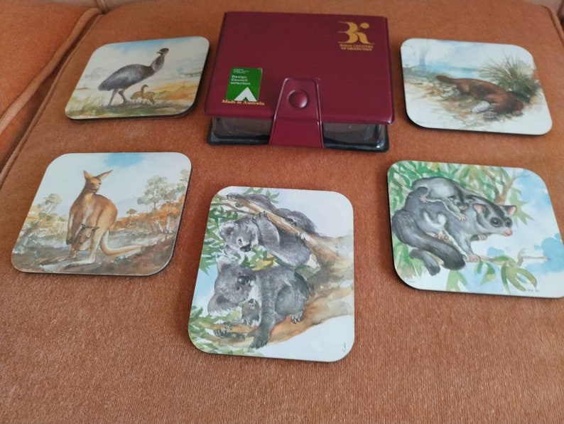 May include: Set of six square coasters featuring Australian animals. The coasters are made of wood and have a glossy finish. Each coaster features a different animal, including a kangaroo, a koala, a possum, an echidna, a platypus, and an emu. The coasters are packaged in a red box with the words "Royal Australian Mint" printed on the front.
