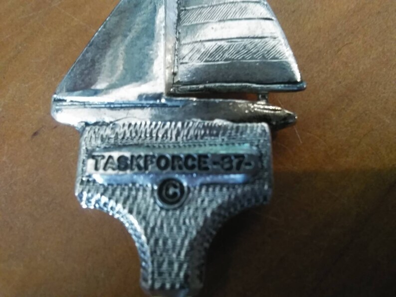 May include: A silver sailboat charm with the inscription "TASKFORCE-37-  &copy;" on the base.