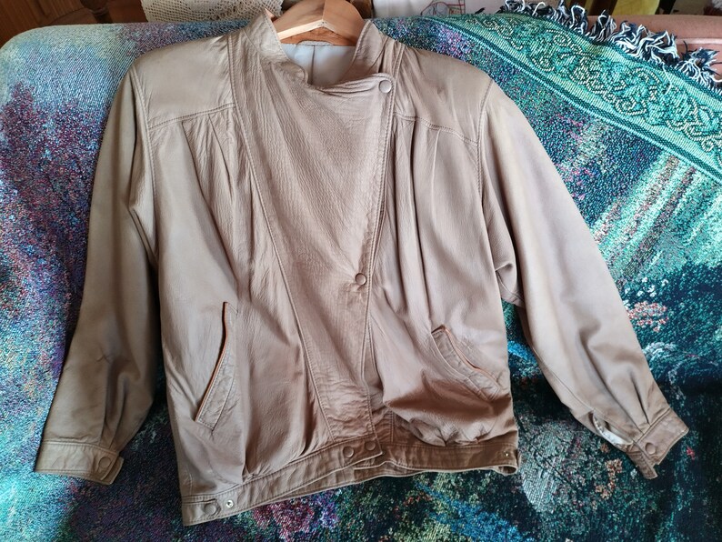 Puede incluir: Una chaqueta de cuero marr&oacute;n claro con cierre de cremallera y un bot&oacute;n a presi&oacute;n en el cuello. La chaqueta tiene dos bolsillos en la parte delantera y una manga larga.