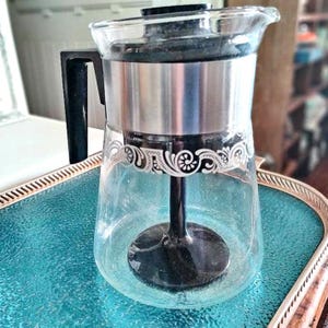 May include: A vintage glass coffee pot with a silver band and a white floral design. The pot has a black plastic handle and a black metal base.