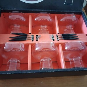 Puede incluir: Un juego de seis vasos de vidrio transparente con asas negras, empaquetados en una caja negra con interior rojo. Los vasos est&aacute;n dispuestos en dos filas de tres, con las asas hacia afuera. La caja est&aacute; abierta, revelando los vasos.