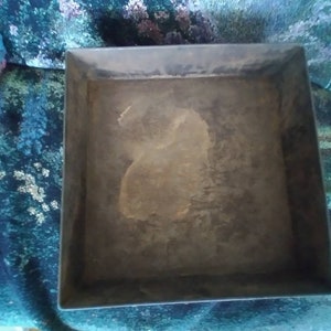 May include: A square metal baking pan with a slightly raised rim. The pan is made of a dark metal and has a worn, rustic appearance. The inside of the pan is empty and shows signs of use.