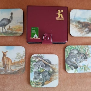 May include: Set of six square coasters featuring Australian animals. The coasters are made of cork and have a glossy finish. The animals depicted are an emu, a platypus, a kangaroo, a koala, and two sugar gliders. The coasters come in a burgundy leatherette case with the text "Royal Charter of Distinction" printed in gold.