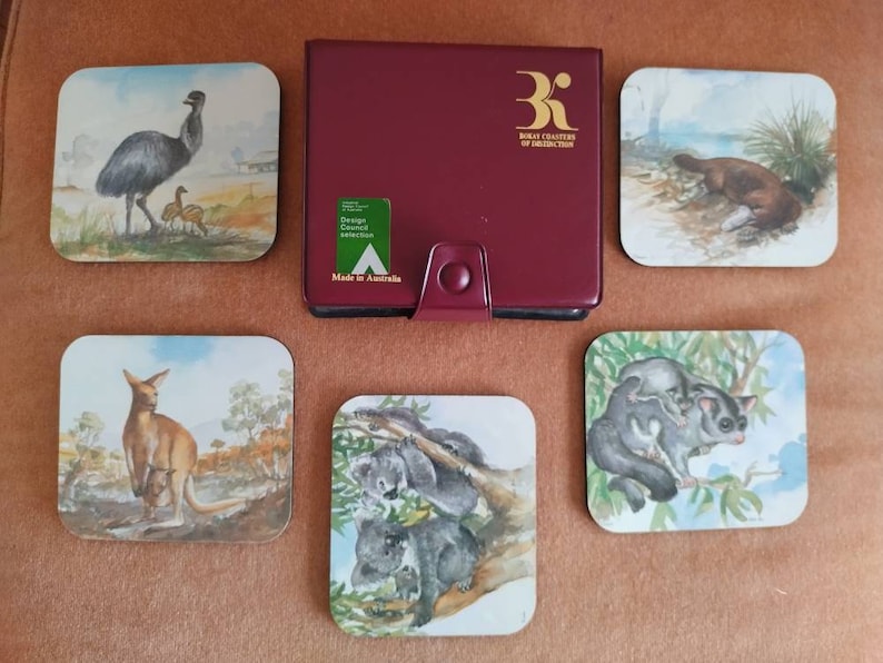May include: Set of six square coasters featuring Australian animals. The coasters are made of cork and have a glossy finish. The animals depicted are an emu, a platypus, a kangaroo, a koala, and two sugar gliders. The coasters come in a burgundy leatherette case with the text "Royal Charter of Distinction" printed in gold.