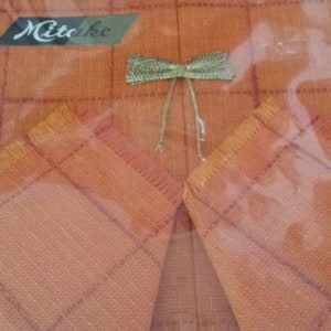 May include: A set of two orange linen napkins with a checkered pattern and fringed edges. The napkins are packaged in a clear plastic sleeve with a green and white border. The package includes a small bow made of gold ribbon. The package is labeled "Mitake".