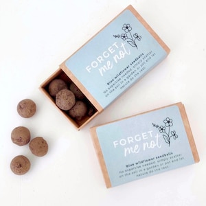 These little brown matchboxes are filled with small round seed balls, spilling out of the box and onto the white table. The boxes have a pretty blue sticker on the front saying 'Forget Me Not' Blue Wildflower Seedballs.