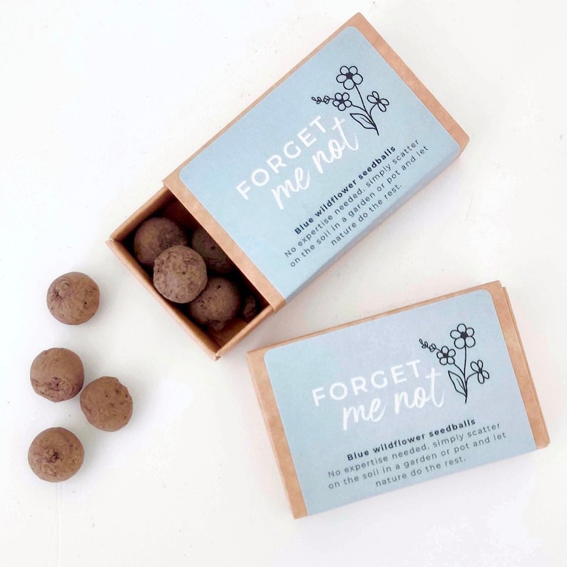 These little brown matchboxes are filled with small round seed balls, spilling out of the box and onto the white table. The boxes have a pretty blue sticker on the front saying 'Forget Me Not' Blue Wildflower Seedballs.