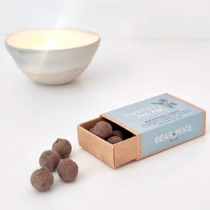 This little brown matchbox with blue label on the top is filled with small brown seed balls spilling out onto the table. In the background we see a cosy lit candle.