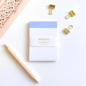 May include: A rectangular notepad with a light blue header featuring the text "TODAY'S LITTLE WINS". A white paper band wraps around the centre with the gold text "BEAR HUGS NOTEPAD 50 SHEETS". Three gold binder clips and a peach pen are positioned nearby.