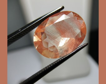 May include: A peach-colored oval gemstone with a shimmery, iridescent finish. The gemstone is held by a pair of metal tweezers.