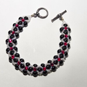 May include: A beaded bracelet featuring black and red beads, with a silver-tone toggle clasp. The bracelet's design incorporates small red beads interspersed among larger black beads, creating a striking contrast. The bracelet is a piece of jewelry.
