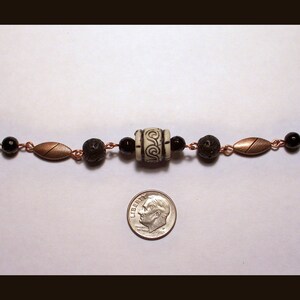 May include: A copper chain necklace with black beads, lava rock beads, and a white ceramic bead with a black swirl design. The necklace is about the size of a dime.