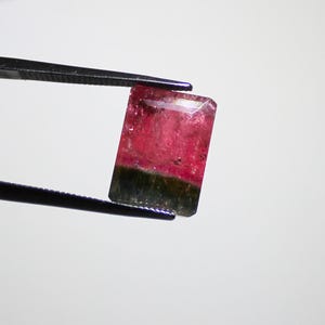 May include: A rectangular watermelon tourmaline gemstone, held by black tweezers. The gem features a vibrant gradient of colors, with a deep pink center transitioning to a green base. The stone is faceted, showcasing its clarity and brilliance.