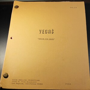 May include: A tan-colored script cover for the TV show "Vegas" with the title "Design for Death" and the production company information: Aaron Spelling Productions, 10201 W. Pico Boulevard, Los Angeles, California 90064. The cover also includes the price $4.50 and the number J-214.