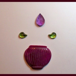 May include: A set of four carved gemstone cabochons. The set includes a purple leaf-shaped cabochon, two green teardrop-shaped cabochons, and a carved ruby-colored cabochon in the shape of a small pot.